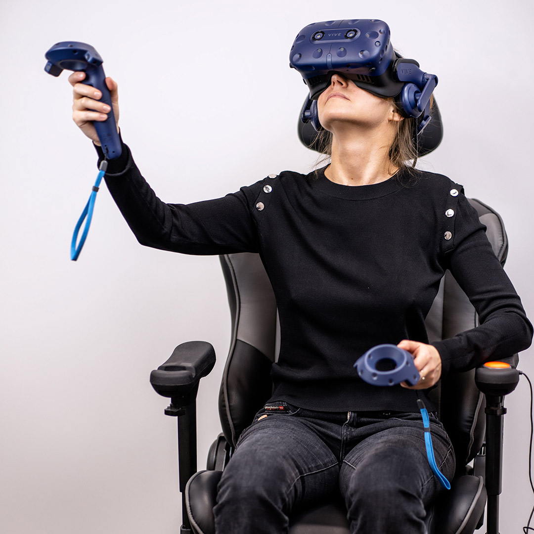 Photo of a woman wearing VR glasses sitting in an office chair, wearing black clothes, using both hands to use blue VR controllers. Left arm is at waist level, right arm is raised to head height. There are visible signs of deep immersion in a VR environment.